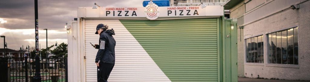 A man in a black jacket walks past a closed pizza shop, looking at his phone, in front of a shuttered storefront with "Hand-Made Fresh Pizza" signage.