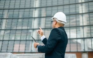 A commercial property manager wearing a white hard hat and suit inspects a modern glass building while holding a tablet, pointing towards the structure.