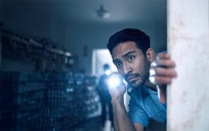 A security guard cautiously peers around a doorway, holding a flashlight that illuminates the dimly lit warehouse. In the background, another figure with a flashlight approaches, adding tension to the scene. The image represents real-time threat detection and the importance of proactive security measures in preventing unauthorized access.