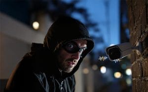 A masked intruder in a black hoodie is caught in the act at night, looking startled as a security camera mounted on a brick wall issues a live audio warning. The camera features sound waves indicating real-time voice intervention, demonstrating the power of Live Audio Voice Down in deterring crime before it happens.