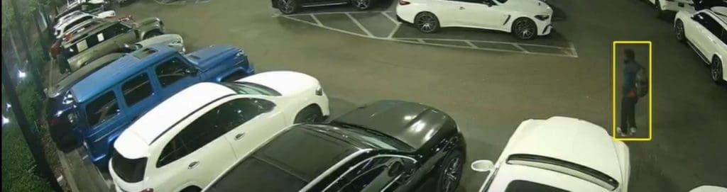  A nighttime security camera feed captures a parking lot filled with vehicles. An AI-powered surveillance system highlights a person standing near a white car with a yellow detection box. The individual appears to be carrying a backpack and wearing dark clothing. The AI system's object recognition helps differentiate between routine activity and potential security threats, reducing false alarms and improving response times.