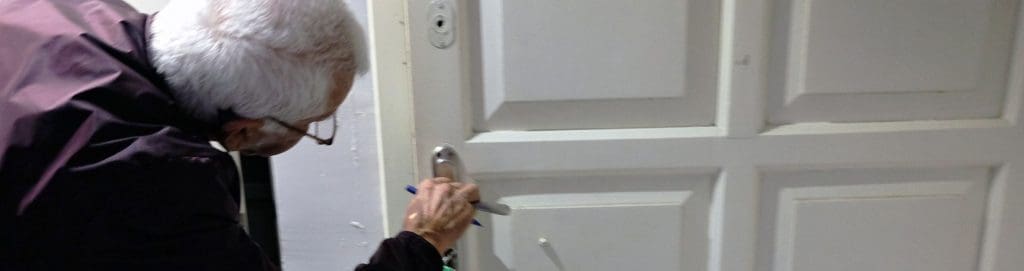 An elderly person with white hair and glasses is inspecting or attempting to unlock a worn-out white door using a tool. The door’s handle and lock appear aged, with visible signs of wear and tear. This image represents the security risks of outdated locks in multi-family properties, emphasizing vulnerabilities like lock picking, unauthorized access, and the need for modern security upgrades such as smart locks and AI-powered live video monitoring.