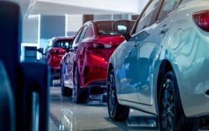 A close-up view of a dealership service lane featuring multiple parked cars, including a white sedan in the foreground and a red car further down the aisle. The brightly lit, modern showroom reflects the sleek design and polished surfaces of the vehicles, representing the professional and high-tech atmosphere of a dealership embracing AI-driven sales and service strategies with EyeQ RevGen Advisor