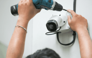 Technician performing security system maintenance by adjusting and securing a wall-mounted surveillance camera with a power drill.