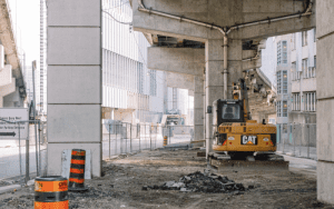 Urban construction site under a highway overpass with heavy equipment and safety fencing, highlighting the need for jobsite security monitoring.