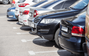 Row of parked cars in a paved parking lot during daytime, showing rear and side views of various modern vehicles lined up in marked spaces.