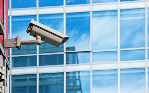 Security camera mounted on the exterior of a modern glass office building, angled for surveillance of the surrounding area.