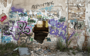 Graffiti-covered, crumbling wall of an abandoned building with a large hole revealing a dark, damaged interior, symbolizing vandalism and urban decay.