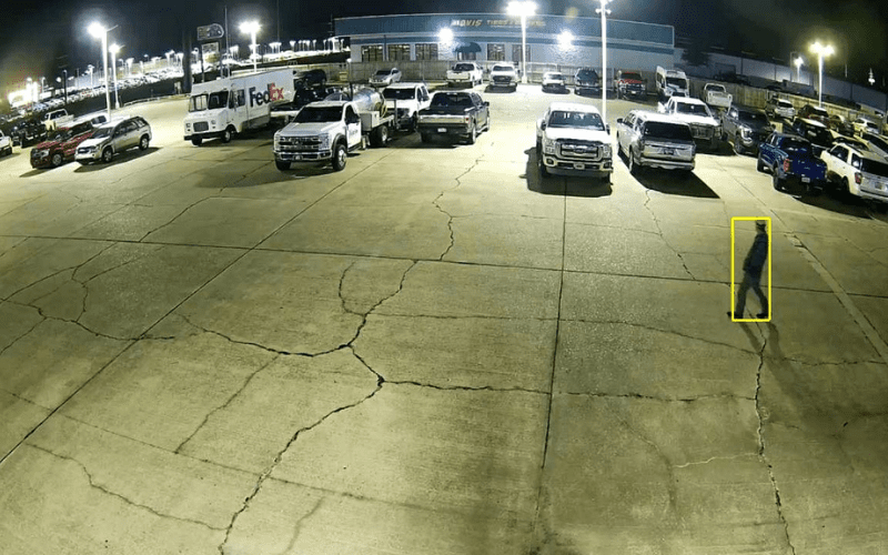 Nighttime surveillance image of an automotive dealership lot with multiple parked vehicles, showing a person walking and highlighted by a yellow detection box to illustrate smart camera monitoring.