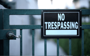 A metal gate with a prominent black and white "No Trespassing" sign mounted on it, partially blocking a narrow walkway between buildings.
