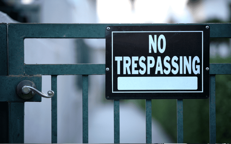 A metal gate with a prominent black and white "No Trespassing" sign mounted on it, partially blocking a narrow walkway between buildings.