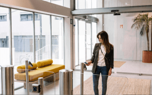 A woman in business attire uses a key card at an access control gate in a modern building lobby with glass walls and a yellow bench nearby.
