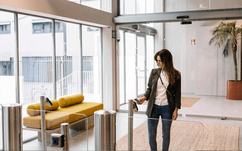 A woman in business attire uses a key card at an access control gate in a modern building lobby with glass walls and a yellow bench nearby.