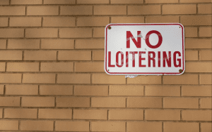 A "NO LOITERING" sign with bold red letters is mounted on a tan brick wall. The sign appears slightly weathered and has minor tape residue or damage near the center. The background consists entirely of evenly spaced horizontal bricks.