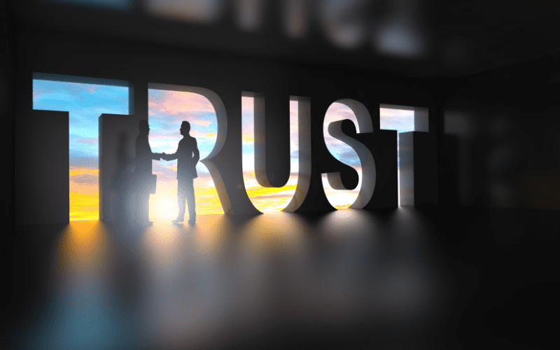 The word “TRUST” in large 3D letters with a glowing sunrise behind it, silhouetting two people shaking hands inside the letters, symbolizing security, partnership, and reliability.