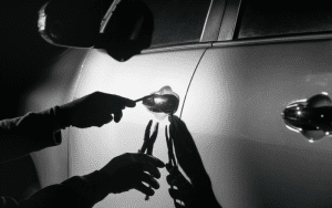 Silhouetted hands using tools to break into a car door at night, highlighting the act of vehicle theft.