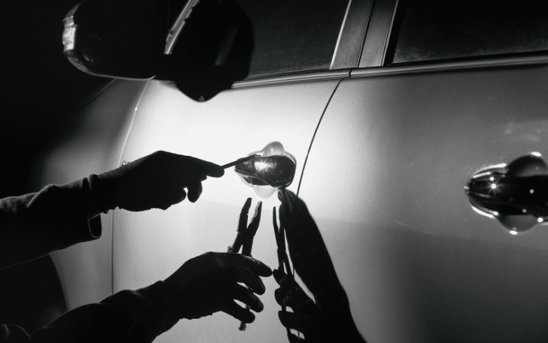 Silhouetted hands using tools to break into a car door at night, highlighting the act of vehicle theft.