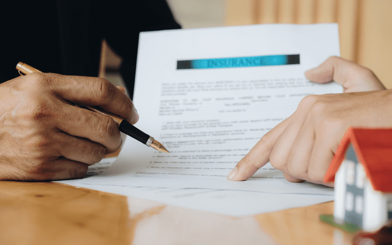 Close-up of two people reviewing and signing an insurance document at a desk, with a small house model in the foreground.