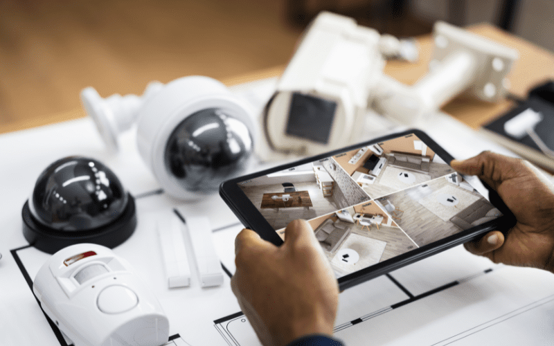 Person holding a tablet showing live security camera feeds, surrounded by various surveillance equipment on a table, symbolizing a modern, integrated security system for multifamily properties.