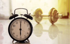 Classic black alarm clock showing 7 o'clock in the foreground with a blurred dumbbell in the background, symbolizing timing and strength in decision-making.