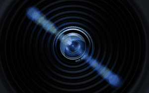 Close-up view of a black security camera lens with a glowing blue flare across the frame, radiating concentric circles to emphasize focus and precision.