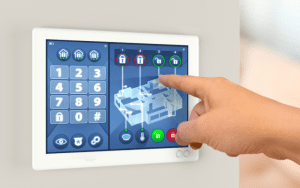A close-up of a hand interacting with a modern touchscreen home security system. The display shows a digital floorplan with locked and unlocked zones, numeric keypad, and control icons for alarms, lighting, and temperature