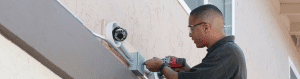 Technician installing a dome-style security camera on the exterior of a building using a power drill, with conduit wiring visible and protective eyewear worn.
