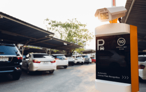 An automatic license plate recognition camera mounted on a post in a covered parking lot. A row of parked cars is visible in the background under a metal canopy, with a sign below the camera displaying 'P', speed limit, and system information."