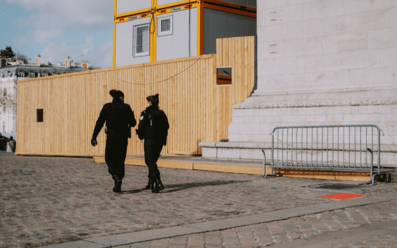 Two uniformed security guards walking past a construction barrier near a large stone structure, illustrating traditional on-site guard presence in urban environments.