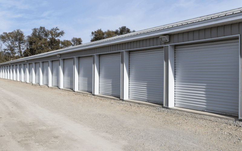A row of closed, white roll-up doors at an outdoor mini storage facility. The units are part of a long metal building with gravel ground in front and security lighting installed above.
