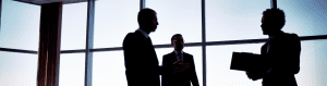 Silhouettes of three business professionals in discussion inside a modern office with large floor-to-ceiling windows, suggesting a formal decision-making setting.