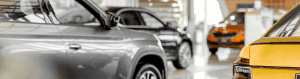 Close-up view of multiple parked vehicles inside a modern auto dealership showroom, featuring a mix of gray, black, and yellow cars under bright lighting.