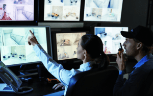 Two security operators in a dark control room monitoring multiple live CCTV camera feeds on large screens, one pointing at a screen and the other speaking into a walkie-talkie.