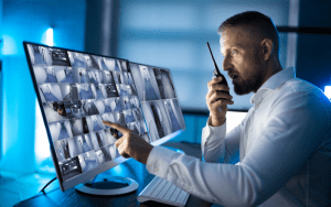 Security professional monitoring multiple live surveillance camera feeds on a dual-screen setup, speaking into a walkie-talkie in a dimly lit control room environment.