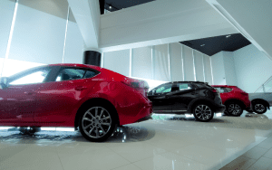 Interior of a modern car dealership showroom featuring several new vehicles lined up in a row, including red and black SUVs and sedans, under bright natural lighting with floor-to-ceiling windows.