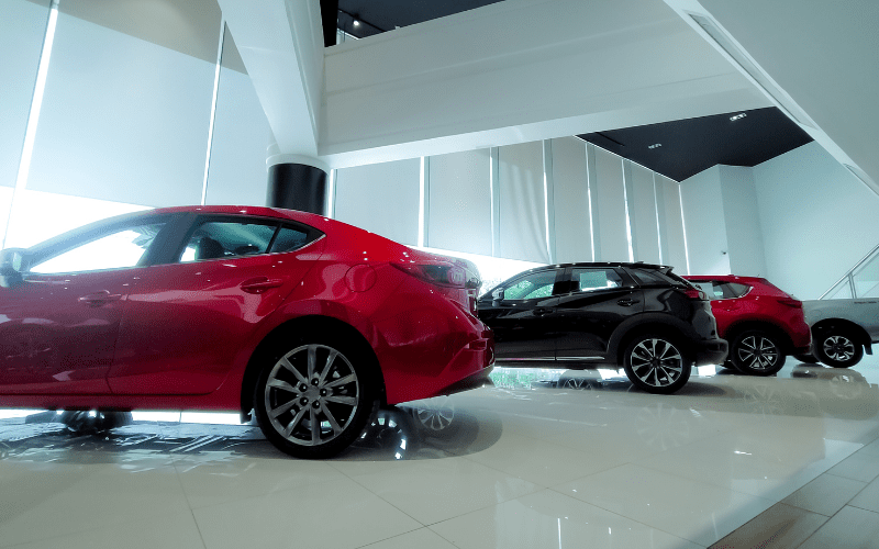 Interior of a modern car dealership showroom featuring several new vehicles lined up in a row, including red and black SUVs and sedans, under bright natural lighting with floor-to-ceiling windows.