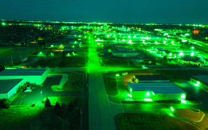 Aerial nighttime view of a large commercial area illuminated by bright green lighting, showcasing industrial buildings, roads, and parking lots under night vision camera effect.
