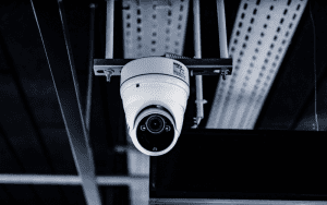 Close-up of a ceiling-mounted dome-style NVR security camera with visible wiring and metal support brackets, set in an industrial or commercial building interior.