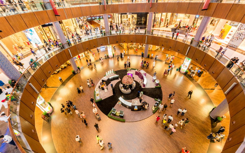 Top-down view of a multi-level shopping mall with crowds of shoppers walking across the central atrium and upper floors, showcasing retail activity and open public space design.