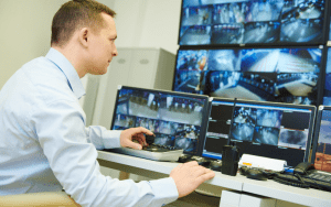 Security professional operating a multi-screen workstation in a control room, actively reviewing live camera feeds as part of a monitoring and surveillance system.
