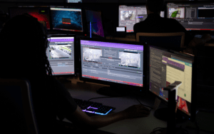 Person monitoring a CCTV system in a dark control room, surrounded by multiple screens displaying live security footage, alerts, and communication tools.